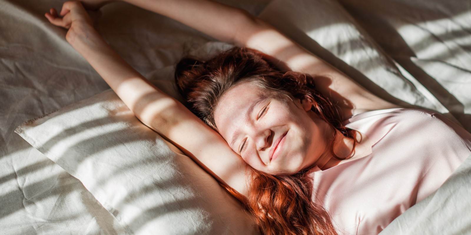 Frau mit roten Haaren streckt sich im Bett nach einer scheinbar erholsamen Nacht.