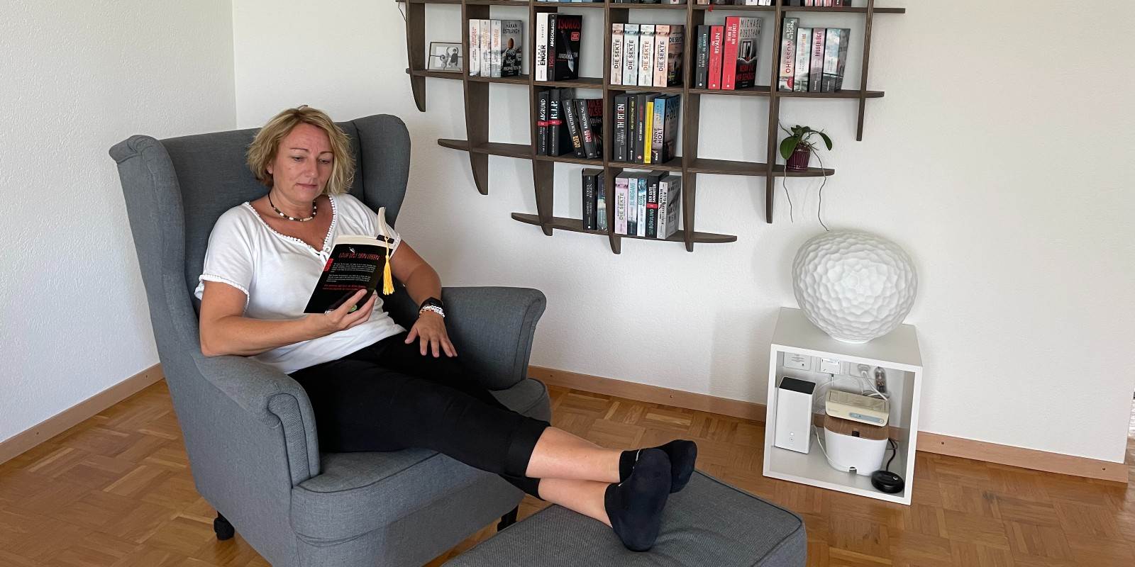 Frau am Lesen eines Thriller-Romans in einem Sessel vor ihrem Bücherregal voll mit Thriller-Romanen.